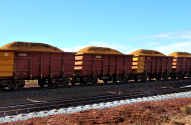 Vagões de trem carregados de minério em trilhos sob céu azul. Terreno avermelhado.