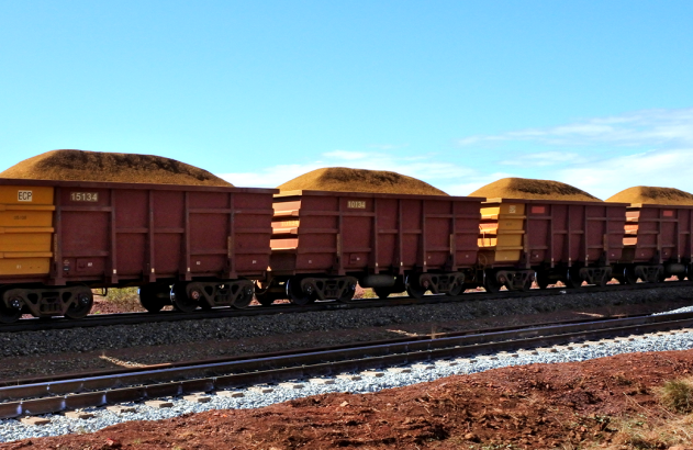 Vagões de trem carregados de minério em trilhos sob céu azul. Terreno avermelhado.