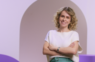 Mulher loira sorridente, braços cruzados, fundo roxo com formas geométricas.