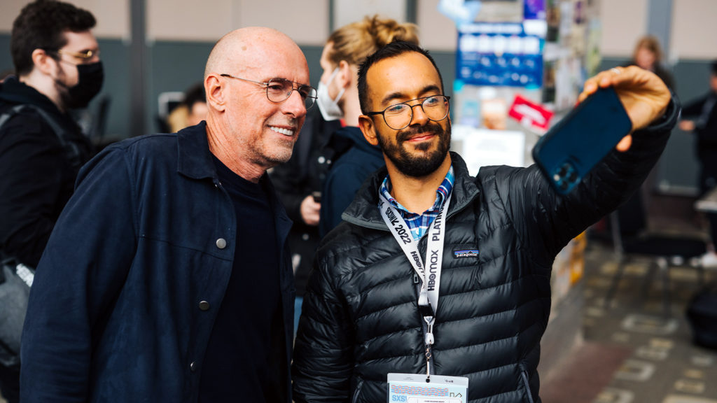 Dois homens, um jovem com celular, tiram selfie com outro mais velho, sorrindo, em evento SXSW.