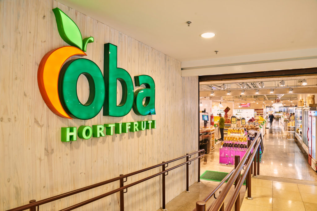 Placa "Oba Hortifruti" em madeira e rampa de acesso a uma loja iluminada com produtos frescos e clientes.