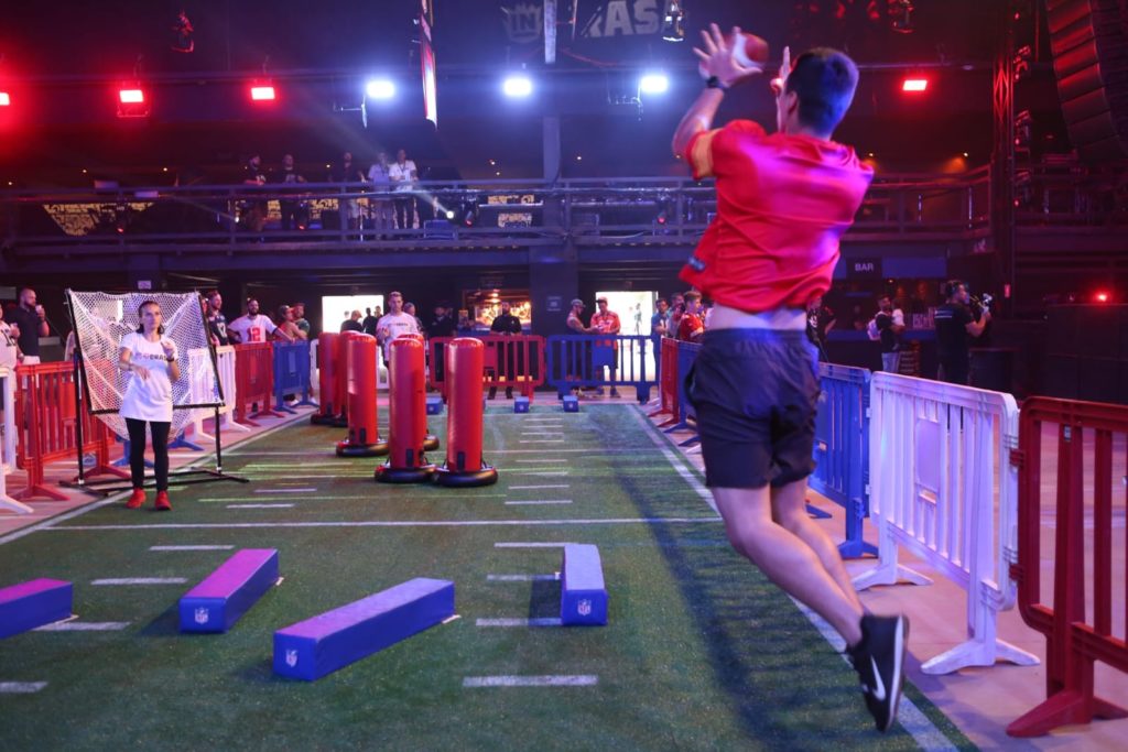 Homem salta para pegar bola de futebol americano em percurso indoor com obstáculos.
