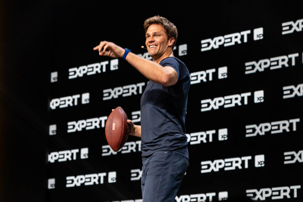 Tom Brady sorrindo em palco, apontando e segurando uma bola de futebol americano. Logotipos "EXPERT XP" no fundo.