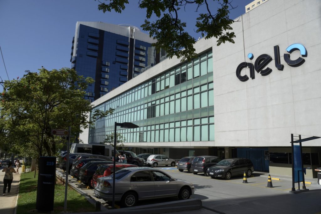 Edifício da Cielo com logo, estacionamento com carros e pessoas, e prédio espelhado ao fundo.