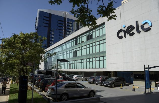 Edifício da Cielo com logo, estacionamento com carros e pessoas, e prédio espelhado ao fundo.