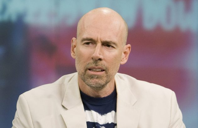 Homem careca e barbudo em casaco claro, olhando para a esquerda. Fundo desfocado com tela colorida.