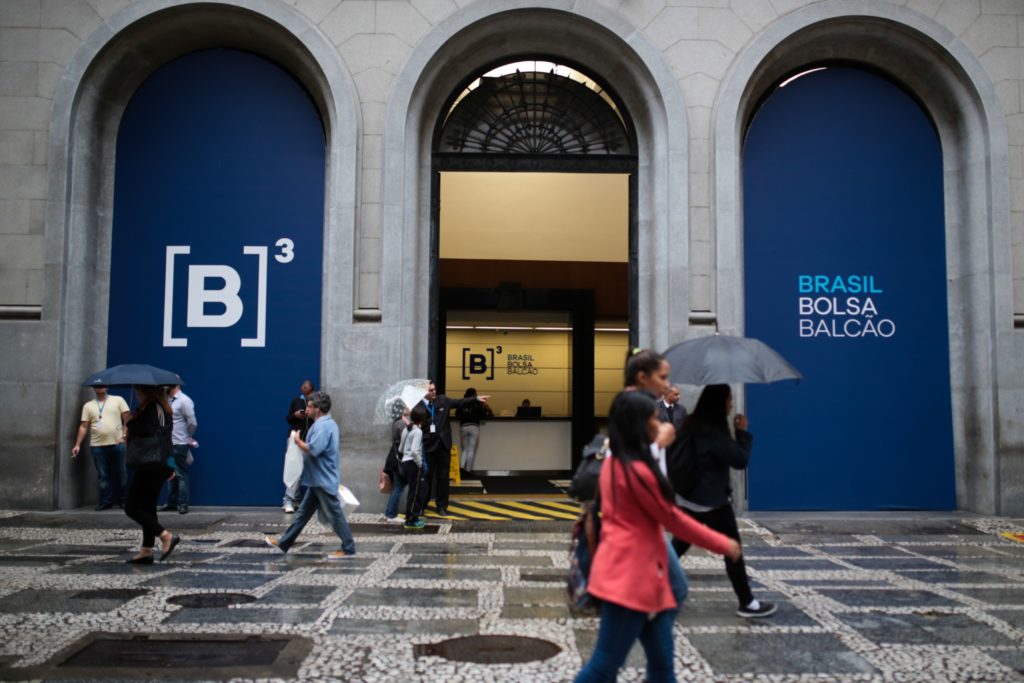 Pessoas andam na rua molhada em frente ao prédio da B3 (Bolsa de Valores do Brasil), sob arcos. Alguns com guarda-chuva.