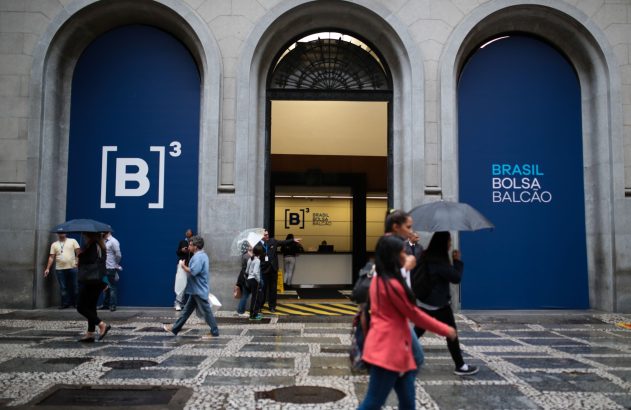 Pessoas andam na rua molhada em frente ao prédio da B3 (Bolsa de Valores do Brasil), sob arcos. Alguns com guarda-chuva.