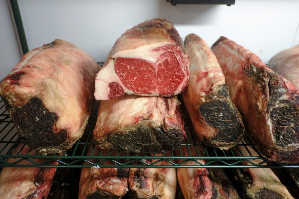 Cortes de carne bovina em maturação a seco, um deles cortado, em prateleiras metálicas.