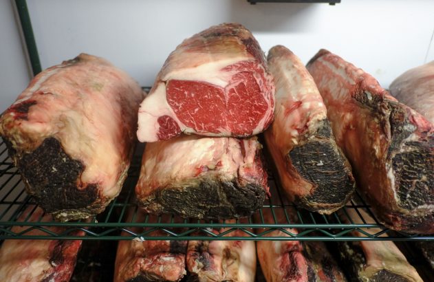 Grandes cortes de carne dry-aged em prateleiras. Uma peça está cortada, mostrando interior vermelho marmorizado.