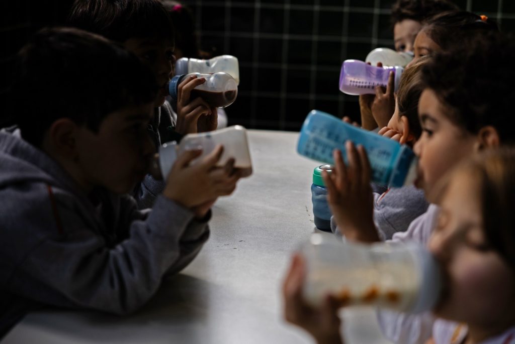 Várias crianças sentadas à mesa bebem líquidos em mamadeiras e copos. O ambiente é escuro.