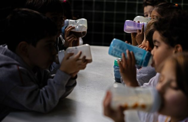 Várias crianças sentadas à mesa bebem líquidos em mamadeiras e copos. O ambiente é escuro.