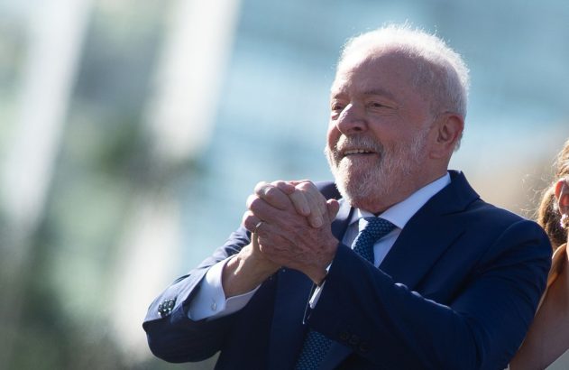 Homem de barba branca e terno sorrindo, mãos unidas. Fundo claro e desfocado.