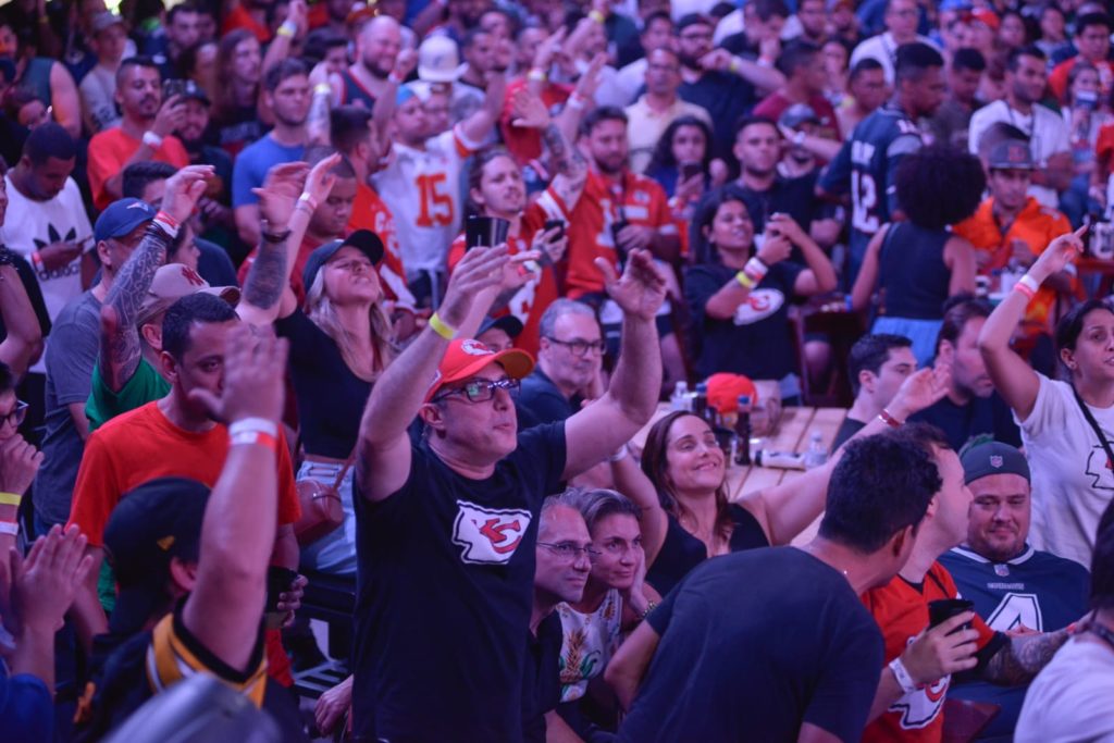 Torcida vibrante com as mãos para cima, muitos em camisas de futebol americano, assistindo a um jogo.
