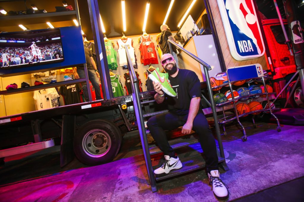 Homem barbudo em óculos escuros segura um tênis verde neon, sentado em escadas de um display móvel da NBA.