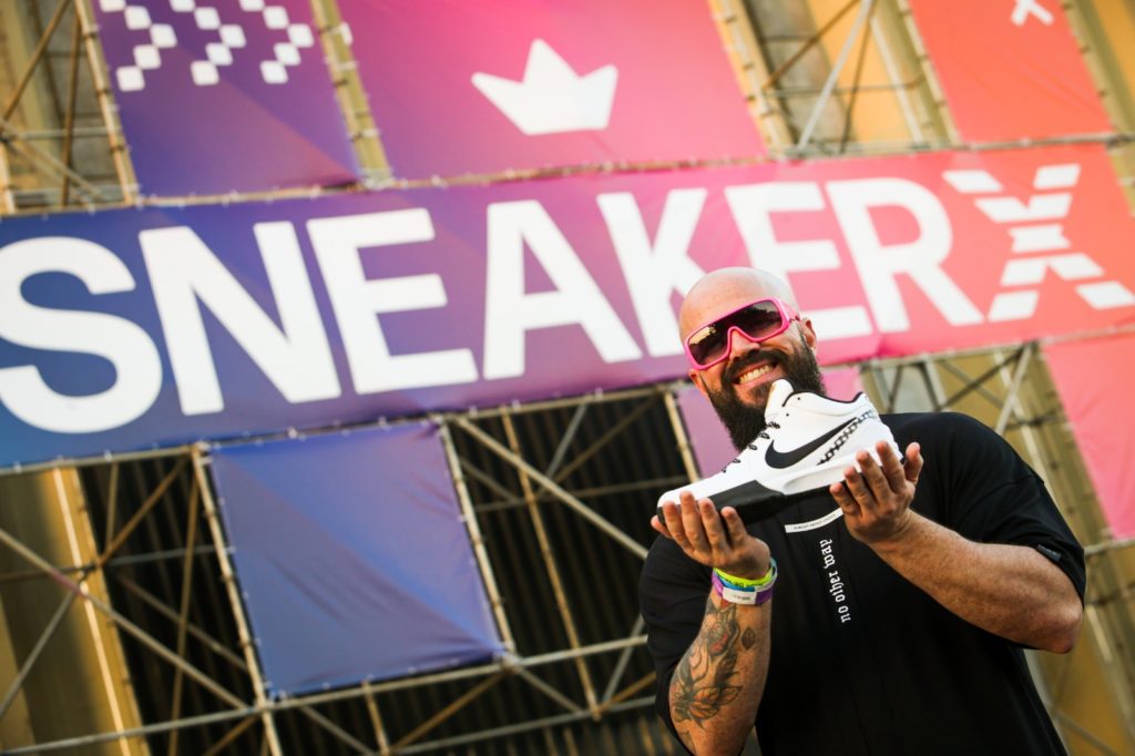 Homem barbudo sorridente de óculos rosa segura tênis branco e preto Nike. Fundo: banner "SNEAKERX".