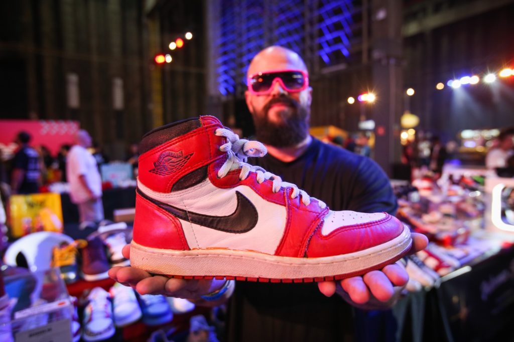 Homem careca de óculos rosa exibe tênis Air Jordan 1 vermelho, branco e preto. Fundo de evento.