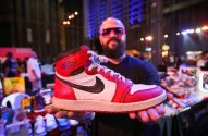 Homem careca de óculos rosa exibe tênis Air Jordan 1 vermelho, branco e preto. Fundo de evento.