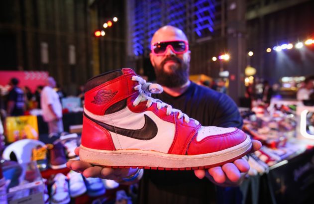 Homem careca de óculos rosa exibe tênis Air Jordan 1 vermelho, branco e preto. Fundo de evento.