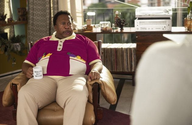 Homem em poltrona, camisa rosa com frutas, segurando caneca "WORLD'S BEST RETIREE". Ambiente interno.