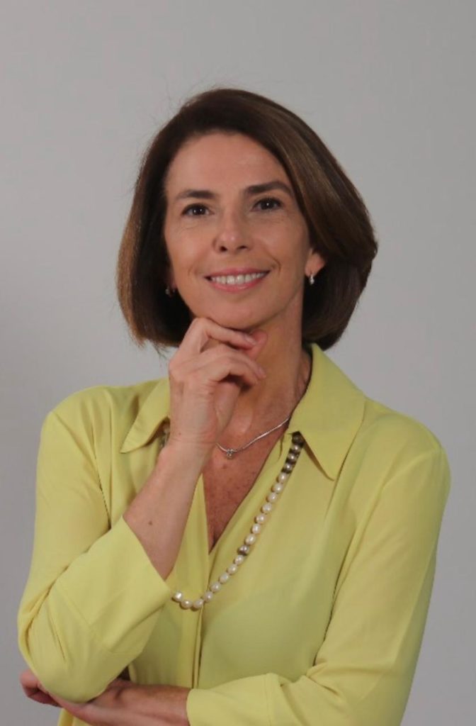 Retrato de mulher sorrindo, mão sob o queixo. Cabelo curto e blusa clara.