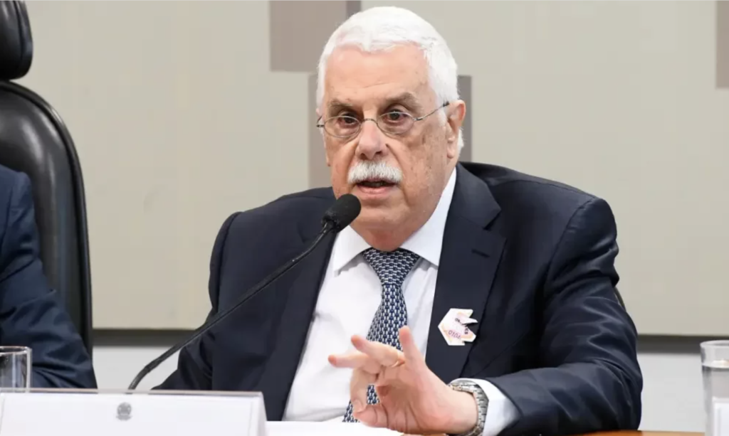 Homem idoso de terno e óculos, com bigode branco, fala ao microfone. Ele gesticula e usa um broche na lapela.