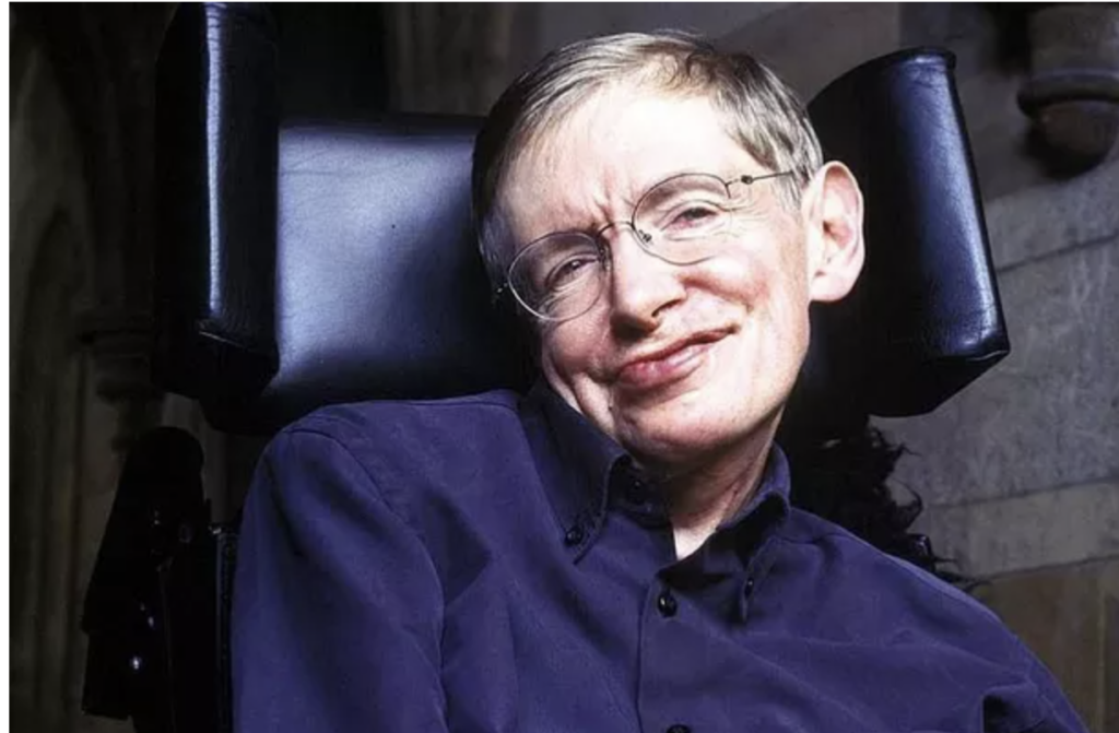 Stephen Hawking, de óculos e camisa azul, sorrindo em sua cadeira de rodas.