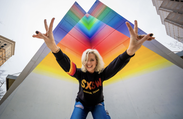 Mulher sorridente posa com "V" de vitória em frente à parede colorida do SXSW.