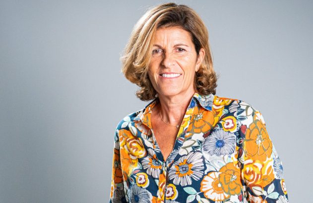 Mulher sorridente com cabelo castanho e camisa floral colorida, olhando para a frente em fundo cinza.