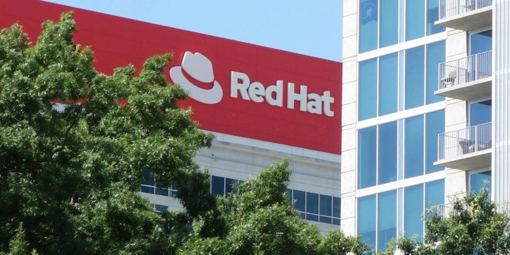 Prédio da Red Hat com logotipo vermelho, parcialmente visível atrás de árvores, ao lado de edifício moderno.