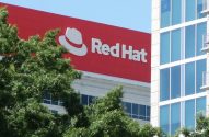 Prédio da Red Hat com logotipo vermelho, parcialmente visível atrás de árvores, ao lado de edifício moderno.
