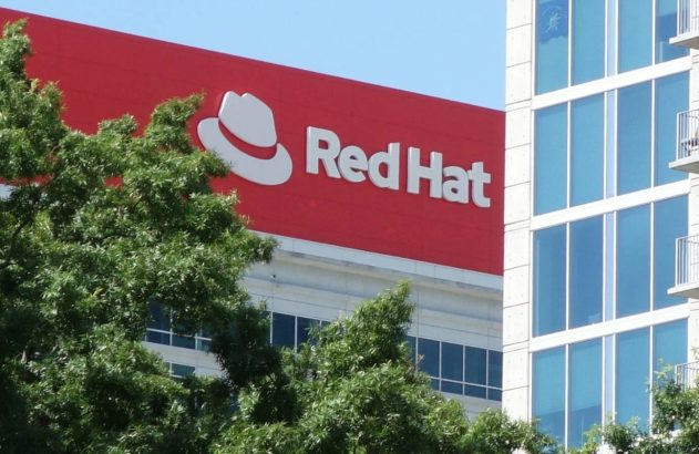 Prédio da Red Hat com logotipo vermelho, parcialmente visível atrás de árvores, ao lado de edifício moderno.