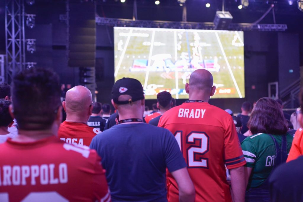Pessoas assistem a jogo de futebol americano em telão. Alguns usam camisas de times.