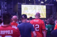 Pessoas assistem a jogo de futebol americano em telão. Alguns usam camisas de times.