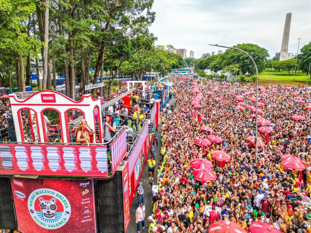 Multidão com guarda-chuvas vermelhos em bloco de Carnaval, seguindo carro alegórico em rua.