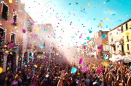 Multidão celebrando em rua com confetes coloridos caindo. Prédios históricos ao fundo.