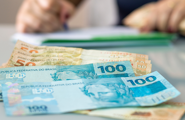 Cédulas de reais (R$100 e R$50) sobre uma mesa, com pessoa escrevendo ao fundo.