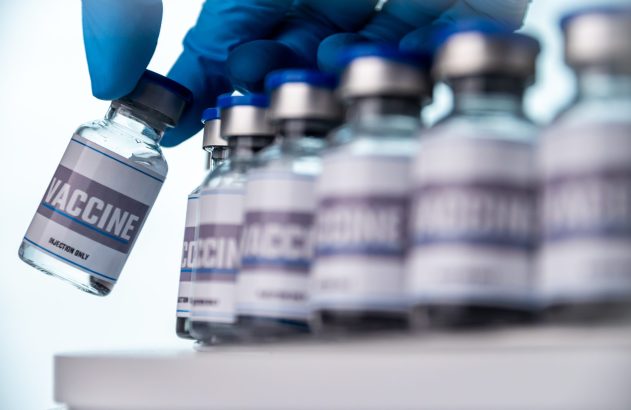 Mão enluvada pega frasco de vacina rotulado 'VACCINE' de uma fileira. 'INJECTION ONLY' também visível.