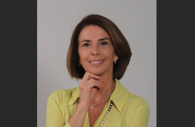 Mulher sorrindo, com a mão no queixo e camisa verde-clara, contra um fundo liso e claro.