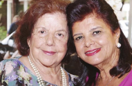 Duas mulheres sorridentes posam próximas para a câmera. A da esquerda, mais velha, usa colar de pérolas.