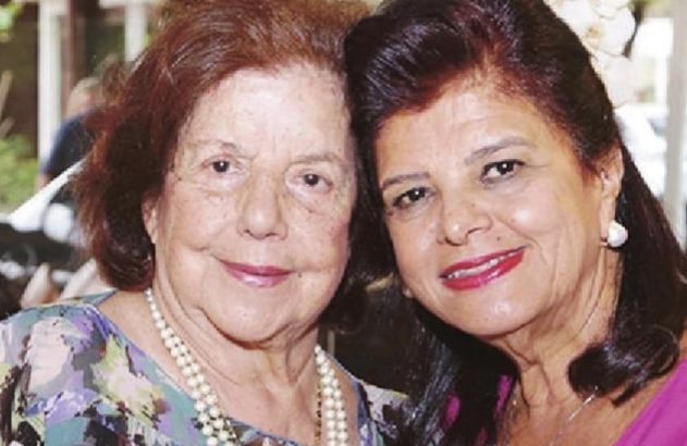 Duas mulheres sorridentes posam próximas para a câmera. A da esquerda, mais velha, usa colar de pérolas.