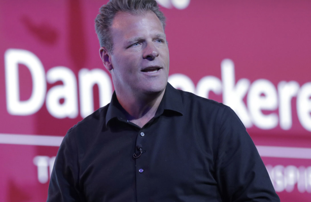 Homem de camisa escura, com microfone, discursando em evento. Fundo com nome "Dan" e "inspire".