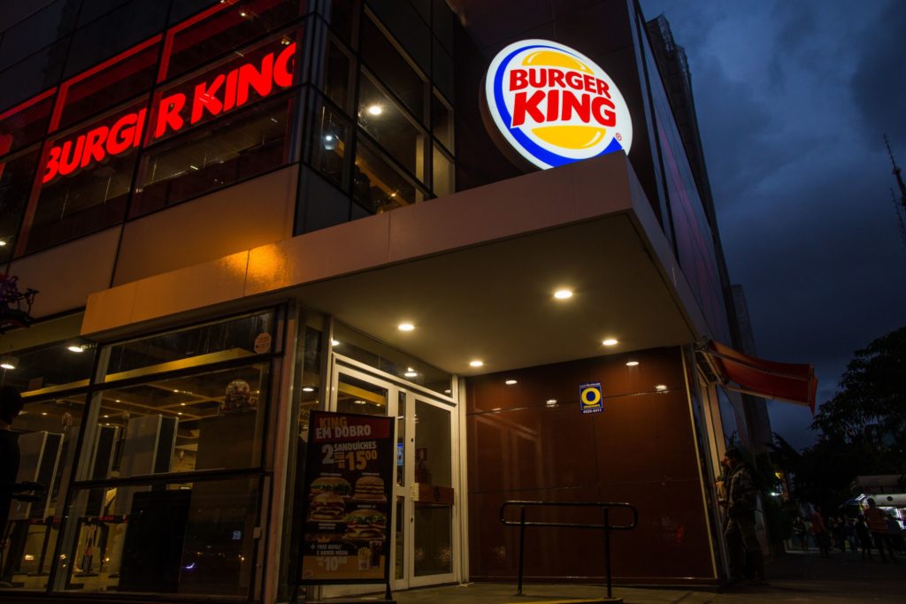 Fachada de um Burger King iluminado ao anoitecer, com seus logos e um cartaz de promoção: 2 sanduíches por R$15.