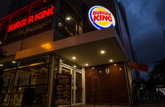 Fachada de um Burger King iluminado ao anoitecer, com seus logos e um cartaz de promoção: 2 sanduíches por R$15.