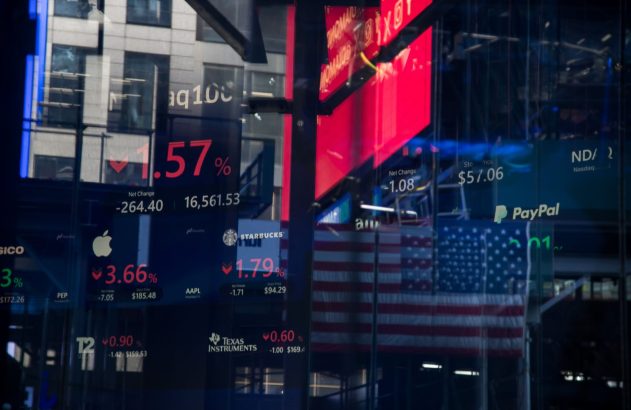 Tela digital exibe dados de ações em queda, com Apple e Starbucks, refletindo edifícios e a bandeira dos EUA.