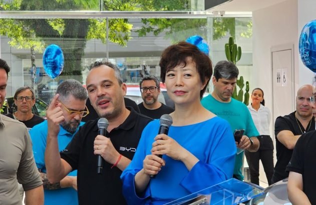 Mulher e homem com microfones falam em evento. Outras pessoas observam.