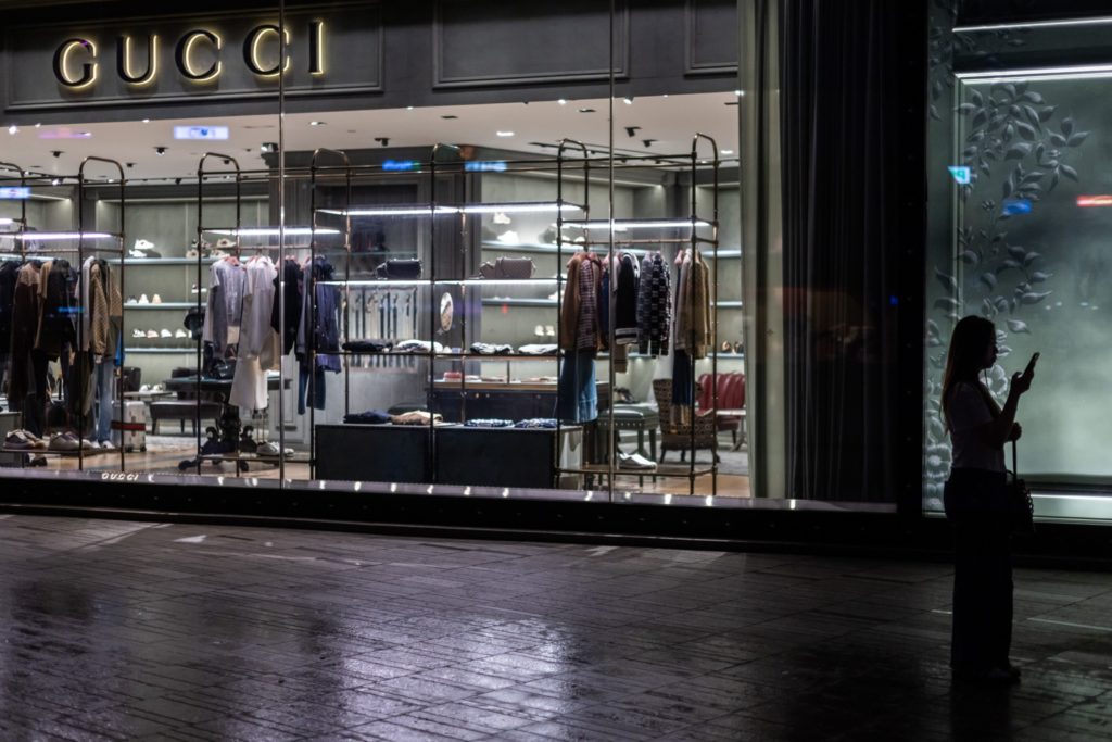 Fachada de loja Gucci à noite, com vitrine iluminada. Uma pessoa em silhueta olha o celular na rua molhada.