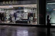 Fachada de loja Gucci à noite, com vitrine iluminada. Uma pessoa em silhueta olha o celular na rua molhada.