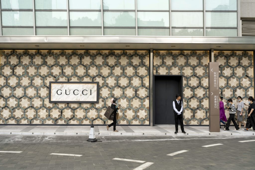 Fachada da loja Gucci com padrão e letreiro. Pessoas andam na calçada, um segurança está de pé.