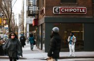 Rua movimentada com o sinal da Chipotle. Pedestres andam na calçada e atravessam na faixa de segurança.